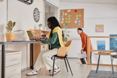 Modern bir ofis ortamında çalışan iki profesyonel, biri masa başında dizüstü bilgisayarla oturuyor ve diğeri de arka planda elinde belgelerle dolaşıyor.