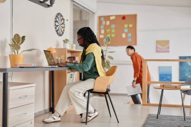 Modern bir ofis ortamında çalışan iki profesyonel, biri masa başında dizüstü bilgisayarla oturuyor ve diğeri de arka planda elinde belgelerle dolaşıyor.