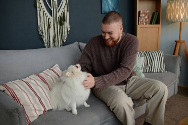 Adam rahat oturma odasında rahat mobilyalar ve modern dekorasyonlar eşliğinde kanepede otururken evcil hayvanla kaliteli vakit geçirmenin keyfini çıkarıyor.