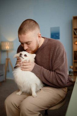 Genç adam rahat bir oturma odasında, küçük, tüylü bir köpeğe sarılıp insan ve evcil hayvan arasındaki bağı gösteren bir sandalyeye oturuyor.