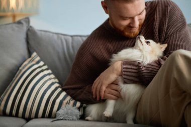 Kanepede oturmuş gülümseyen ve kucağında tüylü bir köpek tutan bir adam. Yanında çizgili bir yastıkla evin içinde rahat bir ortam.