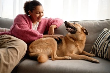 Gülümseyen kadın, Alman çoban köpeğiyle rahat bir kanepede, büyük pencerelerden içeri ışık süzülen parlak, davetkar bir oturma odasında eğleniyor.