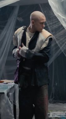 Duvarlarda şeffaf çarşaflarla kaplı atölyedeki aynaya mükemmel uysun diye el yapımı yama ceket deneyen genç beyaz adamın dikey geniş açılı görüntüsü.