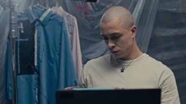 Orta ölçekli, beyaz bir adam, arka planda asılı duran gömlekleri yeniden tasarlamak için şeffaf çarşaf desenlerle kaplı bir şekilde, dizüstü bilgisayarın önünde, resim stüdyosunun önünde oturuyor.