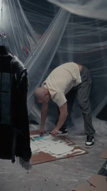 Beyaz tenli genç adam dikey olarak şablonla yere beyaz boya sürüyor. Tekerlekli paten kullanıyor, giysilere boya yapmaya hazırlanıyor. Stüdyoda şeffaf çarşaf duvarlarla çalışıyor.