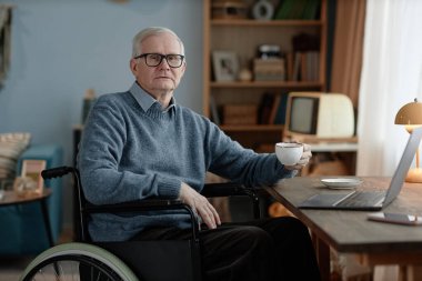 Beyaz saçlı yaşlı erkek tekerlekli sandalye kullanıcısının portresi masadaki çay fincanı ile çekilmiş rahat ev ortamında dizüstü bilgisayarı olan, boşluğu kopyalayan