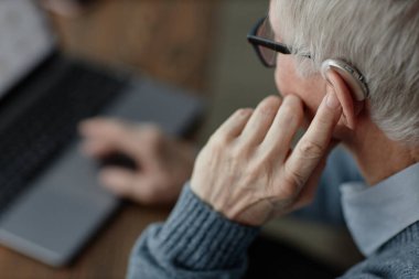 Close up on elderly mans hand tuning up hearing aid behind ear by pressing finger on device controlling volume of sound while working on laptop at home, assistive technology use, copy space clipart