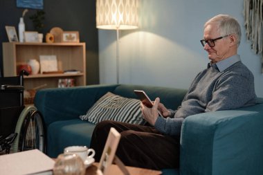 Tekerlekli sandalyede rahat bir oturma odasında rahat bir koltukta otururken dijital tablet okuyan yaşlı erkeklerin yan görüntüsü.
