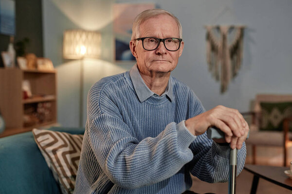 Medium portrait of calm elderly man with glasses wearing blue knitted sweater sitting on couch with walking cane in cozy living room, copy space