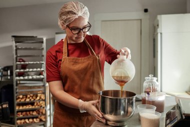 Orta boy kadın fırıncı saç filesi takmış metal kaseye akçaağaç pekmezi dökerken fırın mutfağında zanaatkar ekmeği pişirirken.