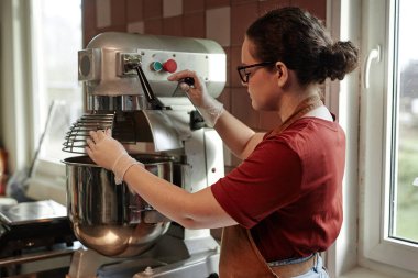 Tek kullanımlık eldiven giyen, büyük yoğurma makinesi kullanan genç bir kadının yan görüntüsü.