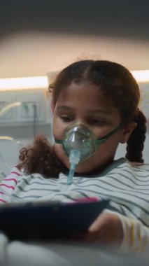 Yüzünde oksijen maskesi olan, koğuşta tedavi görürken konuksever yatağında oturan ve resim çizen küçük hasta Afrikalı Amerikalı kızın dikey görüntüsü.