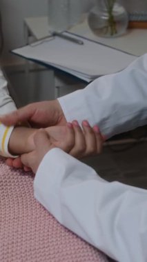 Omuzlarında steteskop olan, beyaz önlüklü, hastanede yatan küçük bir hastanın elini tutup nabzını ölçen, dikey ve tanınmayan bir doktorun görüntüsü.