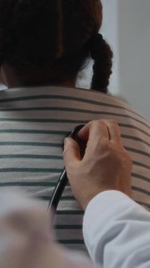 Beyaz önlüklü, tanınmayan bir doktorun dikey görüntüsü. Akciğerlerinin steteskop takıp hastanede oturan küçük kızı dinlemesi.