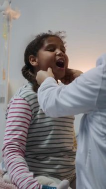 Beyaz önlüklü tanınmayan bir doktor tarafından muayene edilen, hastane koğuşunda lenf düğümlerini avuçlayarak gezen küçük Afrikalı Amerikalı kızın dikey orta pozu.