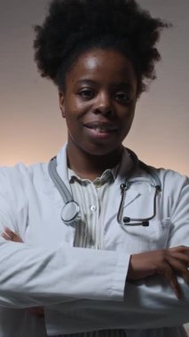 Genç, dost canlısı, omuzlarında steteskop bulunan laboratuvar önlüğü giyen, kolları çaprazlanmış kameraya gülümseyen siyah kadın doktorun dikey yavaş portresi.