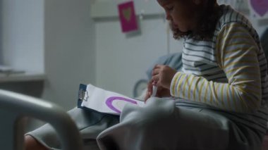 Hastane odasındaki tramvay yatağında pijamalı, duvarında çocuk resimleri olan, kağıt üzerine kalemle gökkuşağı çizen, klinikte tedavi gören küçük siyah kız yavaşlıyor.