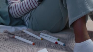 Hasta, küçük, siyah bir kızın, rahat iç çamaşırlı iki kurdelesiyle aydınlık odada battaniyenin üzerine oturmuş, çizimleri tamamlanmış, işaretli, gökkuşağı resmi çizilmiş bir fotoğrafını çek.