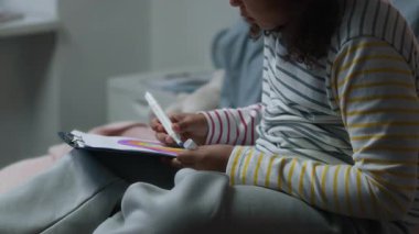 Küçük yetenekli Afro-Amerikan kız rahat pijama giyiyor konuksever yatakta oturuyor ve renkli gökkuşağını kalem kullanarak kağıda çiziyor.