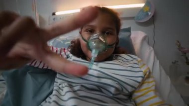 Yüzünde oksijen maskesiyle hastane yatağında yatıp kameraya gülümseyen ve modern klinikte tıbbi yardım alırken barış işareti gösteren neşeli, küçük siyah kızın görüntüsü.