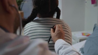 Beyaz önlüklü erkek doktorun akciğerlerinin steteskop kullanarak küçük bir kızı hastane yatağında oturturkenki görüntüsü.
