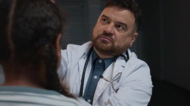 Orta yaşlı, beyaz önlüklü ve omuzlarında steteskop olan beyaz erkek doktorun, klinikteki doktor randevusundaki küçük bayan hastaların kulaklarını incelerkenki yakın çekimi.