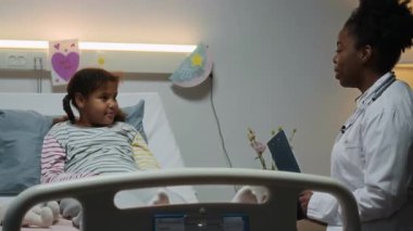 Hasta kartıyla beyaz önlüklü genç siyahi kadın doktor hastane yatağında uzanmış, duvarında çocuk resimleri olan gülümseyen küçük siyah kızla konuşuyor.