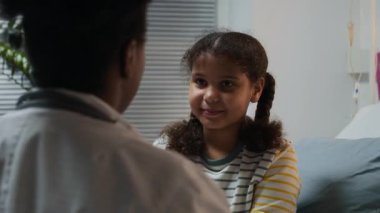 Beyaz önlüklü siyahi kadın boğazını kontrol ederken beyaz önlüklü, açık renkli koğuşta fener kullanan tatlı Afrikalı Amerikalı bir kızın omzunun üstünden.