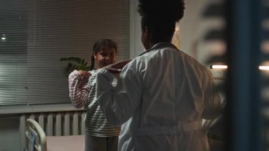Siyahi kadın doktor, beyaz önlüklü. Küçük siyah kız için omuz egzersizi yapıyor. Gece karanlığında hastane koğuşunda tekrarlıyor.