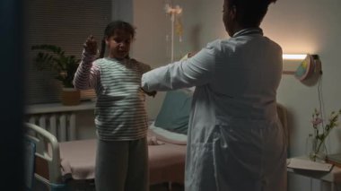 Laboratuvar önlüklü siyahi kadın doktor, küçük siyah kadın hastayla bileklerini ısıtıyor. Arkasından tekrarlıyor. Loş akşam ışığında hastane koğuşunda egzersiz terapisi yapıyor.