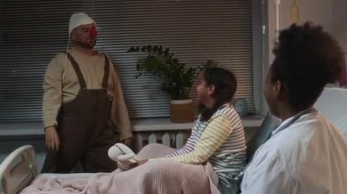 Orta yaşlı komik beyaz erkek palyaço elinde oyuncak olan küçük Afrikalı Amerikalı kızı güldürüyor ve hastane yatağında genç siyahi kadın doktorla koğuşta oturuyor.