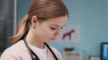 Pembe üniformalı, omuzlarında steteskopla veteriner otoskobu kullanan, modern klinikteki evcil beyaz tavşanın kulaklarını kontrol eden beyaz kadın veteriner pozunu aşağı eğ.