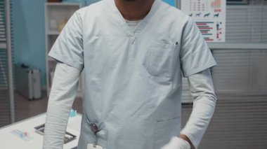 Üniformalı ve steril eldivenli genç Afro-Amerikan erkek veterinerin yavaş yavaş portresini kaldır ve modern veteriner ofisinde hafif kollarını kavuşturup tıbbi hayvan posterleriyle kameraya bak.