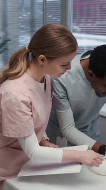 Pembe önlük giyen, elinde hasta kartı olan, genç siyah erkek asistana talimat veren ve klinikteki beyaz evcil tavşanların muayenesinde yardımcı olan beyaz kadın veteriner.
