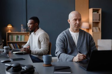 Orta yaşlı erkek iç mimar. Kafası kazınmış bir şekilde dizüstü bilgisayar ekranına odaklanmış bir şekilde çizim yapıyor. Ofisteki meslektaşıyla birlikte masa başında çalışıyor.