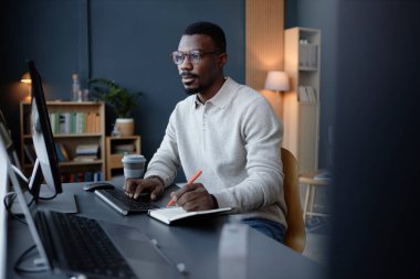 Gözlüklü siyahi adam, mavi duvara karşı yakalanmış, bilgisayar ekranına bakıyor çalışma masasındaki çalışma masasında yazıyor, boşluğu kopyalıyor.