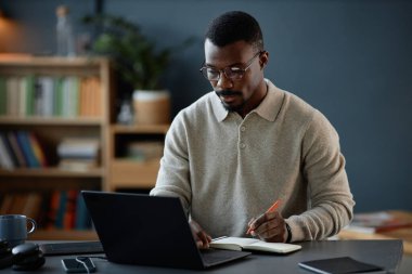 Not defterine gözlük takan yetişkin siyahi bir adamın mavi duvara karşı yakalanmış samimi ofis ofisindeki bilgisayarında çalışırken orta boy fotoğrafı.
