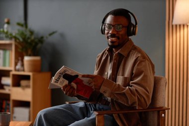 Güzel erkek dergisi okurken kameraya gülümseyen, kulaklıklı, neşeli, yetişkin siyahi bir adamın portresi.