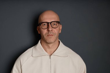 Tıraş olmuş kazak ve gözlük takmış, koyu gri duvara poz veren orta yaşlı, kendinden emin bir adamın stüdyo portresi.