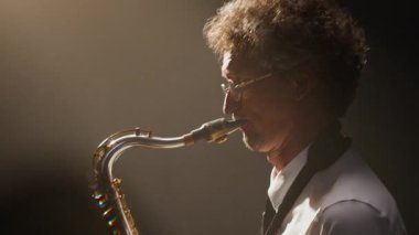 Orta yaşlı, beyaz, klasik kıyafetli erkek müzisyen. Parlak gökkuşağı mercekli saksafon performansı sergiliyor. Enfes enstrümanını koyu arka planda aydınlatıyor.