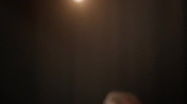 Orta yaşlı, beyaz bir adamın yavaşça kafasını kaldırdığı ve karanlık odada tek başına duran ve yukarıdan gelen ışık huzmeleriyle aydınlatılan kameraya baktığı bir kare.