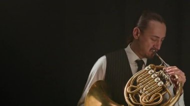 Orta boylu, tutkulu beyaz erkek müzisyen. Klas bir kıyafet içinde. Karanlık sahnede Fransız kornasında nefes kesici bir solo yapıyor.