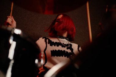 Low angle shot of persistent teenage girl shaking short red hair while playing rhythm on drums during rehearsal session in recording studio with dim light, copy space clipart