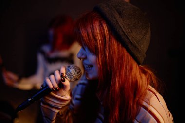 Side view of young female vocalist with red hair singing song devotedly into microphone playing underground punk music with girls band on stage in dim neon light, copy space clipart