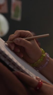 Yurt odasında otururken elinde kalem tutan, defterine resim çizen, tanınmayan genç Z jenerasyonunun orta boy yakın çekimi.