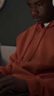 Modern Afrikalı Amerikalı çocuk yurt odasında kanepede oturuyor, kulaklıkla müzik dinliyor ve dizüstü bilgisayarında daktilo kullanıyor.