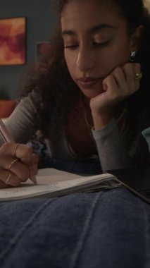 Yurt odasındaki kanepede yatıp dizüstü bilgisayara bakan ve ödevlerini yaparken not alan melez bir kız.