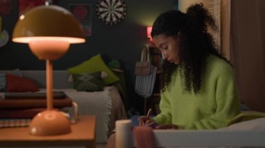 Yurt odasında oturan, yeşil süveter giyen melez genç bir kız not defterine yazıyor.