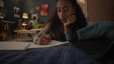 Yurt odasındaki kanepede yatıp dizüstü bilgisayara bakan ve ödevlerini yaparken not alan melez bir kız.