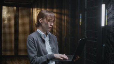 Modern veri merkezindeki rafların sunucusundaki donanım denetimi için dizüstü bilgisayar kullanan beyaz kadın bilişim uzmanının orta boy fotoğrafı.